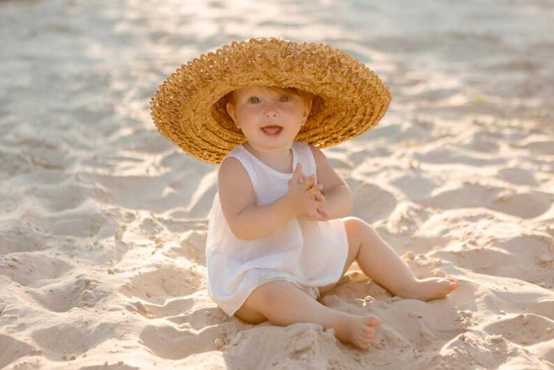 baby girl white clothes straw hat sits white sand beach summer