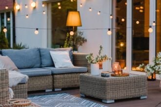 A residential patio with wicker furniture, textiles, and several types of warm lighting, including string lights and candles.