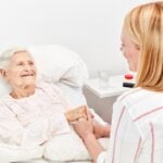 A bedridden elderly woman smiling and holding the hand of her caregiver, who is sitting on the bed's edge.