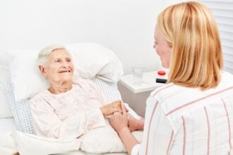 A bedridden elderly woman smiling and holding the hand of her caregiver, who is sitting on the bed's edge.