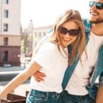 A happy couple outside against a railing. They're smiling with sunglasses on and both are wearing denim with a white top.