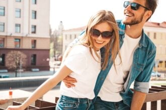 A happy couple outside against a railing. They're smiling with sunglasses on and both are wearing denim with a white top.