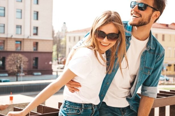 A happy couple outside against a railing. They're smiling with sunglasses on and both are wearing denim with a white top.