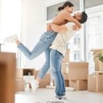 A couple in their 30s standing in the middle of their apartment with packed boxes around them. They're hugging and smiling.