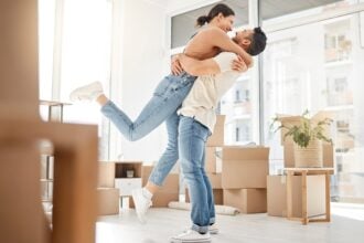 A couple in their 30s standing in the middle of their apartment with packed boxes around them. They're hugging and smiling.