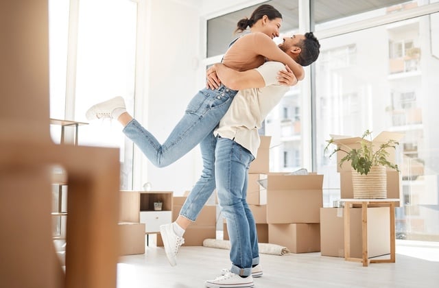A couple in their 30s standing in the middle of their apartment with packed boxes around them. They're hugging and smiling.